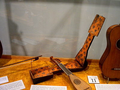 [Mandolin has a light colored wood body with mid-brown color string adjusters. It is laying across its case which is hinged open. There is a dark flower pattern on an orangish background.]